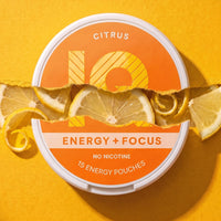 A round tin of IQ Citrus Energy + Focus pouches, shown on a bright yellow background with torn lemon slices and citrus curls layered across the front. The label is orange and white with the 10Q logo and text reading 'CITRUS', 'ENERGY + FOCUS', 'NO NICOTINE', and '15 ENERGY POUCHES'.